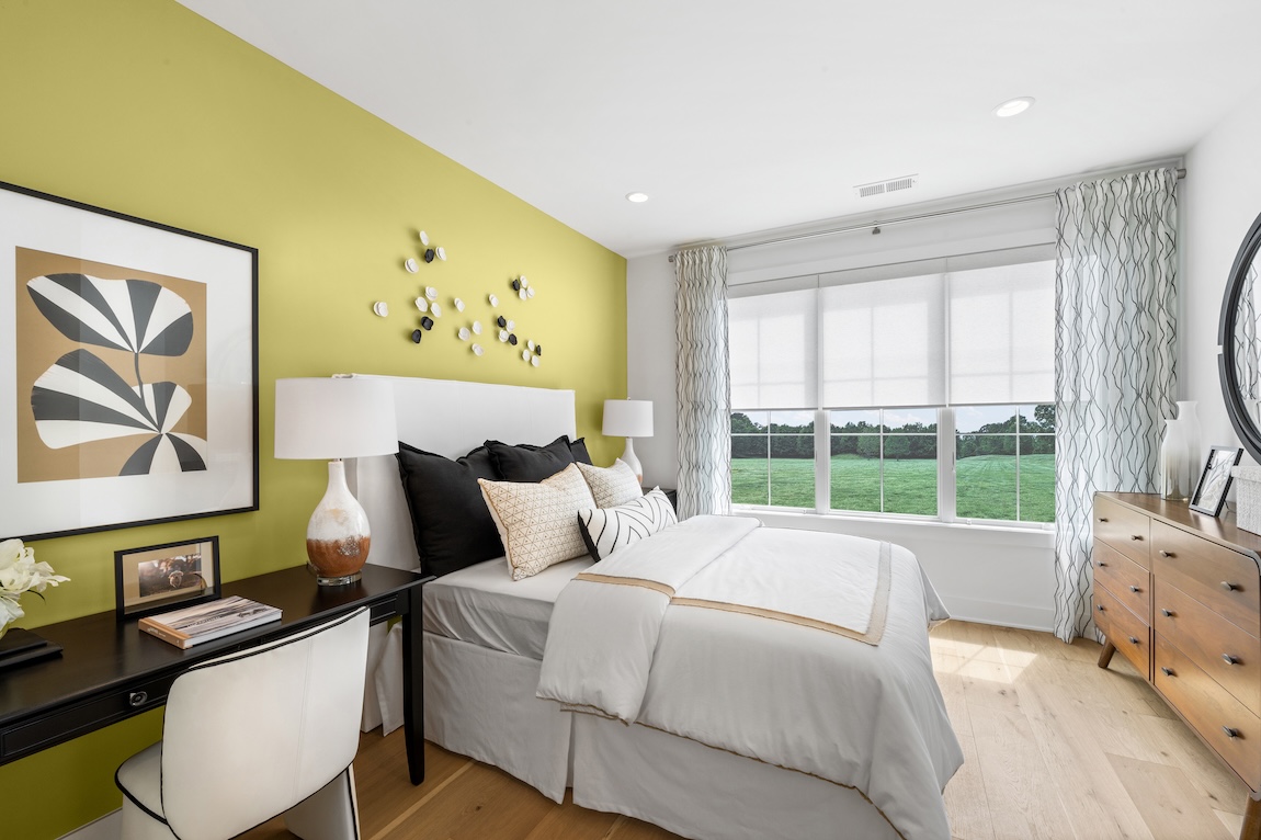 Modern bedroom with lime-green accent wall, monochrome art, and layered neutral bedding in a Toll Brothers home. Natural light floods the space through wide windows overlooking greenery.